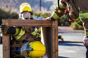 Firefighter crawling through small wooden tunnel, with another firefighter supervising