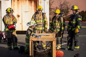 Firefighters gathered around a small wooden tunnel being instructed by another firefighter