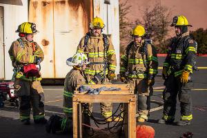 Firefighters gathered around small a wooden tunnel being instructed by another firefighter