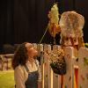 The image features a warmly lit stage setting with two individuals engaged in a whimsical scene. In the foreground, a person with curly hair kneels while looking upward, wearing a light-colored blouse under denim overalls. The second person stands behind a white picket fence adorned with colorful cutouts of butterflies and vegetables. This person is dressed in a vibrant costume, including a yellow outfit with gold tassels and a wide-brimmed hat covered in translucent lace, obscuring their face. They hold a large fabric prop resembling a butterfly chrysalis. Near the fence, there is a decorative bucket filled with artificial flowers. The background is dimly lit, showing a row of empty black chairs.