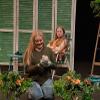 The image shows a stage scene with two young women and various props. In the foreground, one woman is kneeling on the floor, wearing a dark green sweater and blue jeans. She is smiling and holding a small object in both hands, covered by garden gloves. In the background, another woman sits on a striped lounge chair, looking slightly away, with her arms crossed and wearing an orange sleeveless top and denim shorts. Around them are metal buckets filled with green plants and orange flowers. A green wooden screen occupies the background, and a ladder with foliage is partially visible on the right side of the image. The setting suggests an indoor garden or plant-related scene.