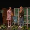 The image shows two individuals on a stage set designed to resemble a garden or outdoor area. On the left, a person is wearing a light pink dress, white shoes, and has a yellow bag over their shoulder. They stand in front of a wooden partition with a green sign hanging from it that reads "TRESSPASSERS WILL BE PROSECUTED." On the right, another person is dressed in a dark green top and blue jeans with white sneakers. They stand in front of a white and green folding screen. Two woven lawn chairs are visible next to the screen, and there is a small side table with a drink on it. In the foreground, at the base of the stage, there are clusters of green foliage with orange flowers.