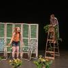 The image depicts a stage performance with two performers. One performer stands in the foreground, wearing an orange top and denim shorts, expressing energetically. Behind them, two blue and white striped folding chairs are placed in front of a wooden screen with green slats. In the background on the right, another performer stands barefoot atop a wooden ladder, draped in a patterned cloth and adorned with foliage. Both the ladder and the base of the stage are decorated with orange flowers and greenery. The setting suggests a natural, outdoor theme.