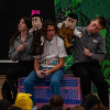 The image depicts a theatrical performance involving three individuals and two puppets. Two performers wearing similar gray shirts are seated on either side of a young person in a white shirt with blue jeans and a championship-style belt over their lap. Each performer on the sides holds a puppet. The puppet on the left is dressed in brown, while the puppet on the right is in a mustard yellow outfit. The backdrop features green foliage patterns. The audience is visible at the bottom of the image, with a child wearing a yellow hood in the foreground.
