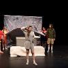 The image shows a theatrical scene with five individuals on stage. In the center, a woman in gray loungewear holds a large notebook, forming a dramatic gesture as if unveiling or revealing something. She stands in front of a rectangular platform covered with a white quilt. Behind her, a backdrop features a collage of hand-drawn images, including numbers, ABC letters, books, and various objects like a basketball and a smiley face. To the left, two people dressed in colorful, casual clothing hold the sheet. On the right, two individuals in jackets observe the scene, one in a green jacket and shorts and the other in a red and white jacket and dark skirt. The stage is dark, focusing attention on the central figures and the backdrop.