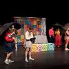 The image depicts a scene on a stage featuring six individuals engaged in a performance. They are positioned in a semi-circle with a colorful backdrop made of a patchwork of squares featuring various smiley face icons in different expressions, such as happy, sad, and surprised. The group is diverse in attire, with a mix of casual and quirky outfits, including bright jackets, skirts, and shorts. Four of the individuals are holding black umbrellas, suggesting a possible thematic element to the piece. In the foreground, a person is focused on a notebook, possibly reading or writing. The stage has a few pastel-colored blocks with smiley faces on them, serving as props.
