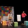 The image depicts a stage scene featuring two people. On the left, a person in casual attire, wearing a beige long-sleeve shirt and shorts, stands with a brown bag. On the right, another person wears a vibrant pink sweater and dark jeans, and is standing with hands on hips. The backdrop consists of a colorful curtain decorated with rows of various emoticons displaying different facial expressions. In front of it, there is a table draped with a light gray cloth. On the stage, there are large cubes with smiley faces painted on them, placed near the individuals. In the center, there's an easel displaying a grid of small emoticons arranged in rows, alongside recognizable icons at the bottom.