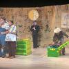 The image shows a theatrical scene set on a stage. In the center, a man and a woman stand together; the man is wearing overalls and a light shirt, while the woman is dressed in a colorful embroidered top. They are sharing a striped blanket wrapped around both of their shoulders. To the right, another person, dressed in a hat and casual clothes, is bending over a green and yellow chest with the lid open. Behind them, a backdrop made of large burlap sacks with various logos and texts is hung. Additionally, a person dressed in black holds a moon-shaped prop on a stick. A yellow and green chest and a yellow crate are onstage.