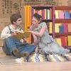 The image depicts a stage scene with two actors seated on the floor. The first actor, wearing denim overalls and a white shirt, is holding a yellow book. The second actor, wearing a light gray dress and a black beret, leans in to look at the book. They are sitting on a multicolored striped rug in front of a vibrant bookshelf filled with colorful books. The backdrop is made of rustic, textured material with indistinct text and logos, giving a textured effect. The setting exudes a playful and theatrical atmosphere with bright, contrasting colors.