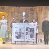 The image depicts a theatrical stage setup with five individuals centered against a backdrop made from stitched, printed burlap coffee sacks. On the far left, a person holding a vibrant yellow umbrella wears a light green dress and black shoes, standing in front of a stack of green and yellow crates. A man in a plaid shirt and beige pants stands with two other individuals holding a large fabric banner. The banner displays black-and-white photos on each side, captioned "Tomás Rivera," with dates "1935-1984." The person in the middle wears an academic gown with a headpiece depicting a photo of the man on the banner. The individual to the right wears a traditional embroidered blouse and dark pants, while the person on the far right is dressed in black. A small colorful rug lies on the wooden floor behind them.