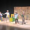 The image shows a group of five performers on a stage. The backdrop consists of burlap sacks with text and logos, suggesting an agricultural theme. The performers are in mid-action, holding props resembling large pieces of corn. From left to right: the first performer is sitting on a stool, playing an acoustic guitar and wearing overalls. The second performer stands, wearing a light-colored shirt and hat, holding a corn prop. The third performer displays a floral blouse and dark pants, also holding corn. The fourth performer, in a plaid shirt and trousers, holds a corn ear. The final performer at the right end sports a black outfit with a hat, also presenting a corn ear. Behind them is a bright green and yellow crate, which is open.