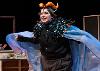 The image shows a person on stage dressed in a theatrical costume. They are wearing a black outfit, covered with a cape made of shimmering blue fabric. The shoulder area is adorned with shiny, sculpted scales in black and orange shades. On their head, they wear a headdress with bright orange horn-like decorations and a small blue ornament on the forehead. The background suggests a theater set, with partial wooden framing behind them.