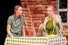 The image captures a theatrical scene featuring two women engaged in conversation. On the left, a young woman with long blond hair styled in braids and wearing a green and white striped t-shirt appears attentive and concerned. She sits at a table draped with a green and white checkered tablecloth. On the right, another woman has long blond hair and wears a green ribbed sweater paired with a light-colored shawl that has fringes. She gestures expressively with her left hand as she speaks. A bunch of yellow bananas rests on the table between them. The backdrop consists of a textured wall resembling brick, enhancing the set's rustic ambiance.