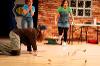 The image depicts a theatrical scene set indoors, characterized by a wooden floor and a brick wall backdrop. Three individuals are prominently featured. On the left side, a person with long hair wearing a brown shirt and camouflage pants is on their hands and knees, reaching towards the floor. Their expression shows concern or urgency as they focus on a few scattered items on the ground. To the right, another individual in a green t-shirt and denim overall dress appears to be moving toward the kneeling figure, with wide eyes conveying surprise or shock. Behind her, a third person, dressed in a light blue shirt and jeans, stands with both hands covering her mouth, also displaying a startled expression. A table is visible in the background, cluttered with various items, while a flag hangs to the left.