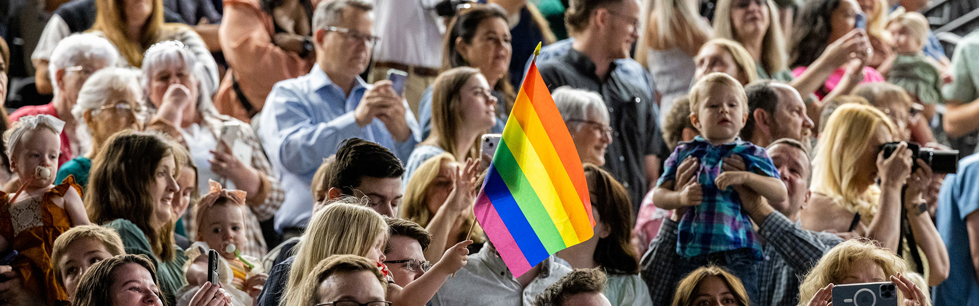 A big crowd with a rainbow flag being held up by a child in the foreground.