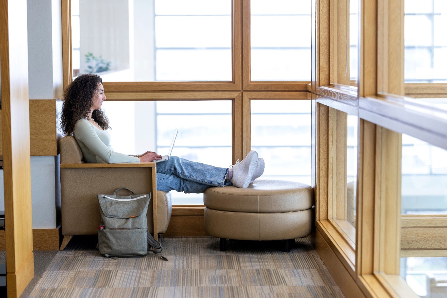 Person working on a laptop while seated in a cozy chair with legs on an ottoman by large windows in the library.