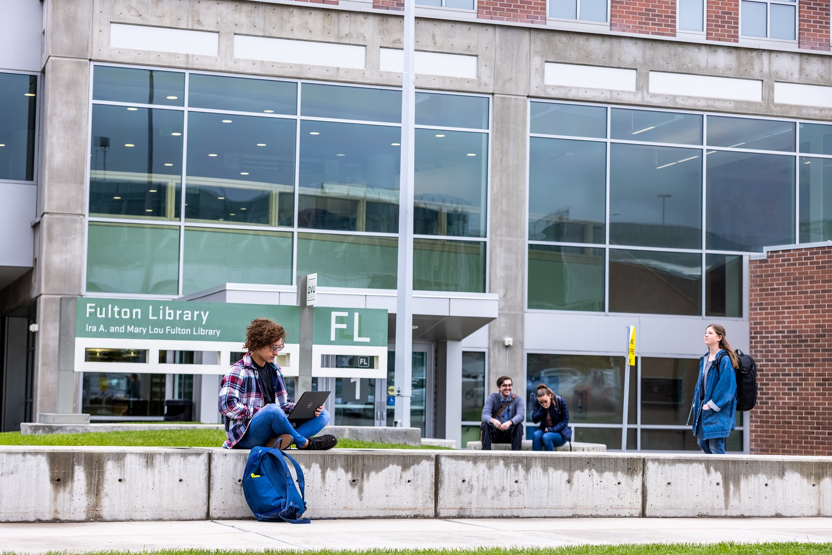 A UVU student sits in front of the Fulton Library with a laptop, while students walk by and mingle in the background.