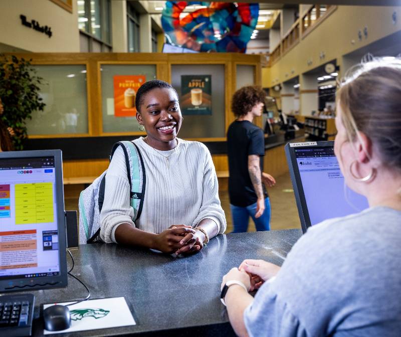 UVU student discussing equipment checkout with a library aide at the circulation desk, with another student walking by in the background.