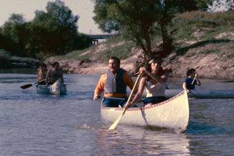 Image of 2 canoes with 2 people in each, rowing down a calm river.