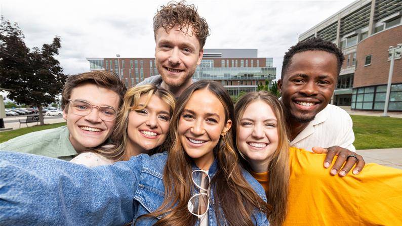 A group of six students pose for a group selfie in front of the Clarke Building on UVU campus.