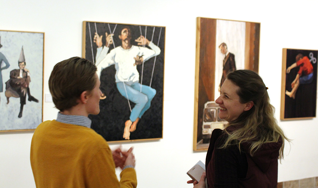 A man and a woman are talking while standing in front of several paintings on a gallery wall
