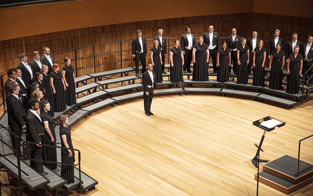 A choir rehearsing in a spacious auditorium with wooden panels and bright ceiling lights.              