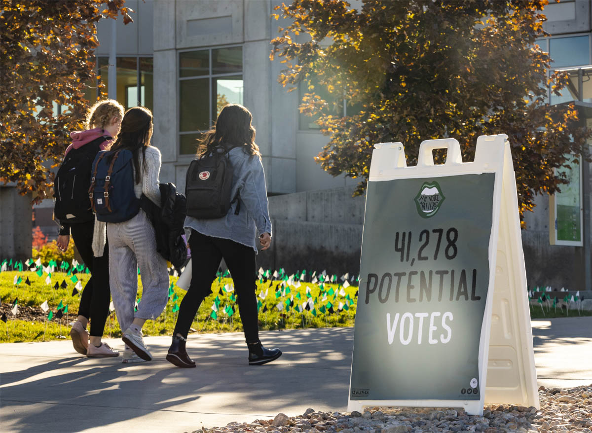 Students pass hundreds of small green, white, and black flags planted in Utah Valley University's lawn in front of the library.