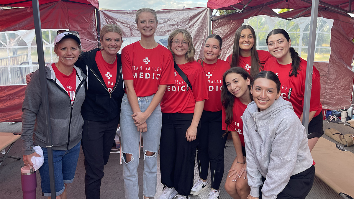 A group of nine students, wearing red "Medic" t-shirts, pose together in a pop-up tent at an event.