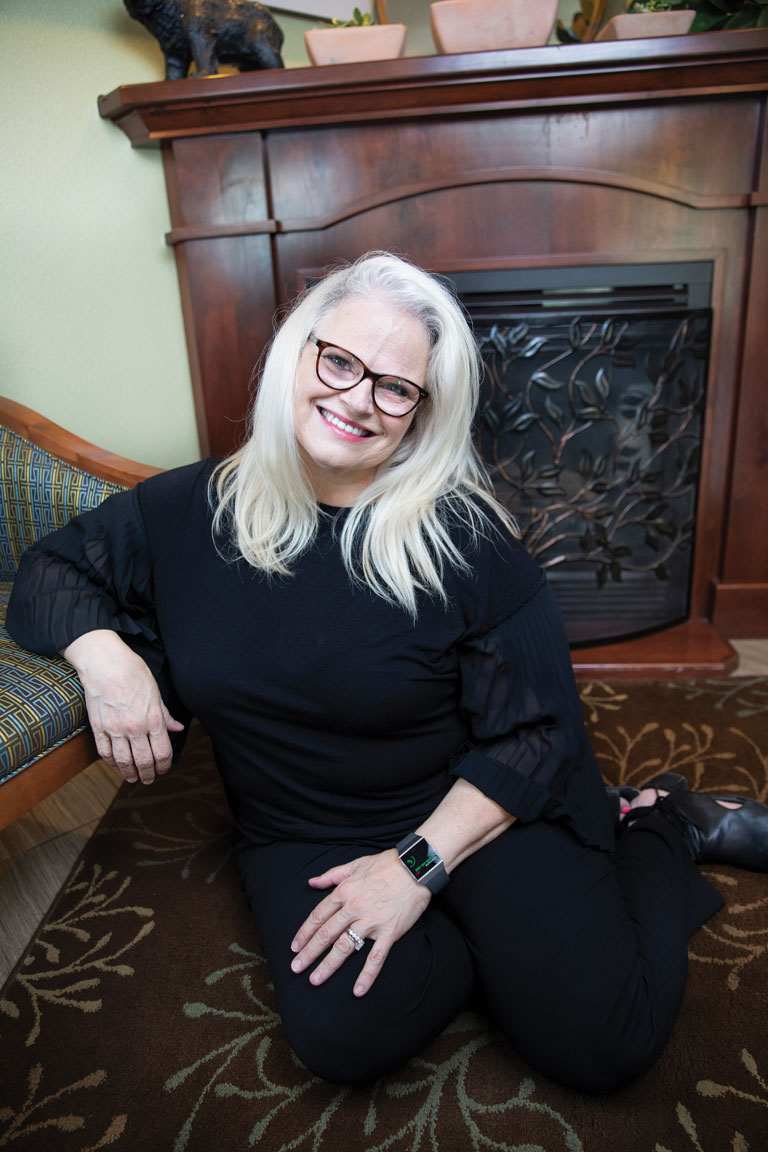 Pamela Neeleman sitting on the floor and leaning on a chair with a fireplace in the background.