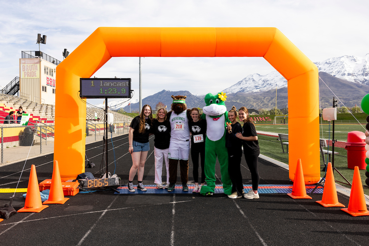 A group of UVU student volunteers and mascots pose for a photo under a bright orange inflatable arch at the 4.01K Race to Retirement. The UVU Wolverine mascot and a green frog mascot stand in the center, with snow-capped mountains visible in the background.