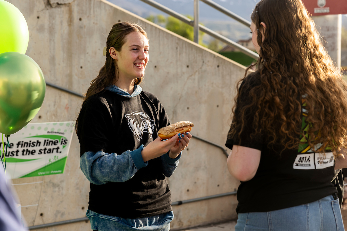 A smiling UVU student volunteer hands a donut to another attendee at the 4.01K Race to Retirement. Green balloons and a sign reading “The finish line is just the start” are visible in the background.