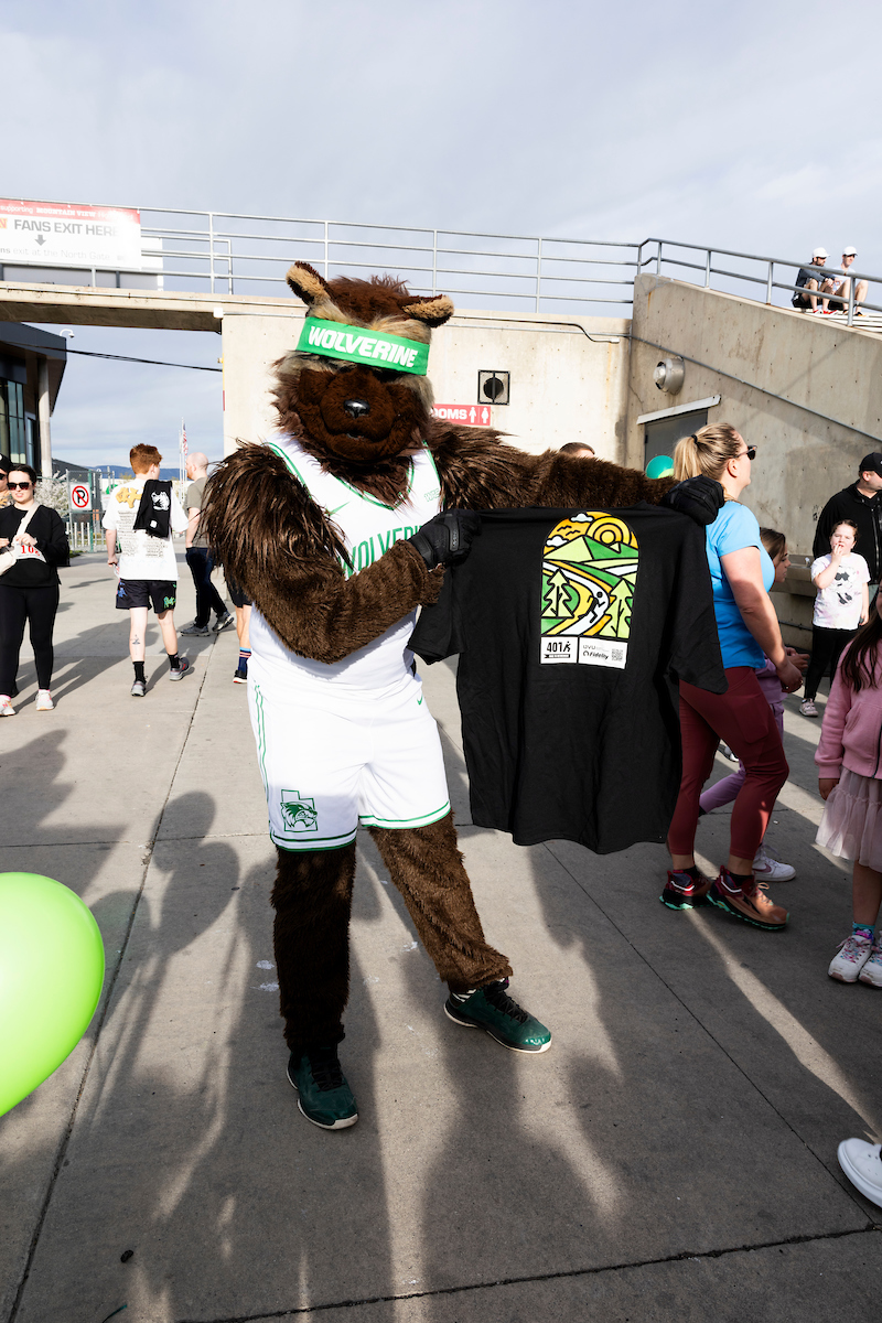 UVU’s Wolverine mascot proudly holds up a 4.01K Race to Retirement T-shirt featuring a colorful mountain and trail design. Participants walk by in the background at the event’s finish area.