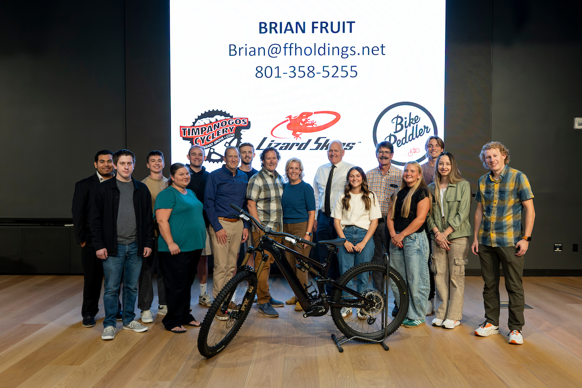 Brian Fruit poses with UVU students, faculty, and staff following his Halladay Lecture, standing in front of logos for Lizard Skins, Bike Peddler, and Timpanogos Cyclery.