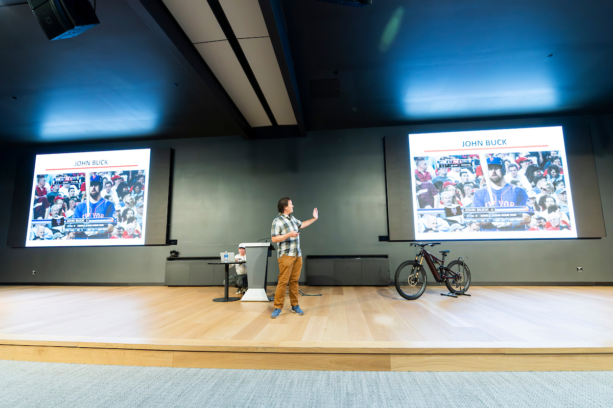 Brian Fruit gestures to a slide featuring former MLB player John Buck while discussing product development and partnerships with Major League Baseball.