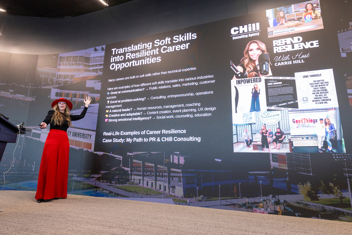 Carrie Hill presents on stage at Utah Valley University with a slide titled "Translating Soft Skills into Resilient Career Opportunities" during the Women in Business Impact Lab event.