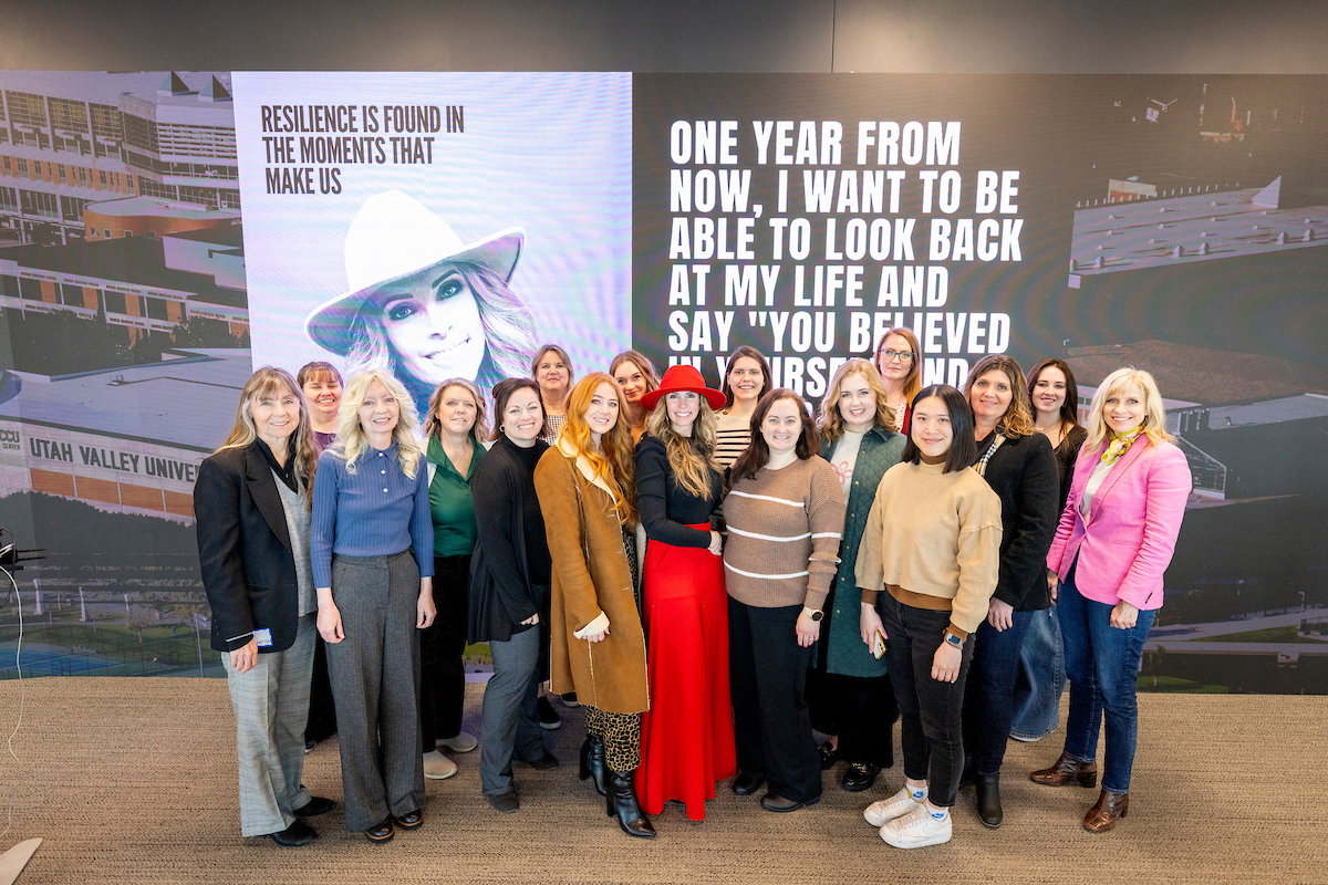 Carrie Hill poses with a group of attendees in front of a large screen displaying her quote, "Resilience is found in the moments that make us," at the UVU Impact DEN event.