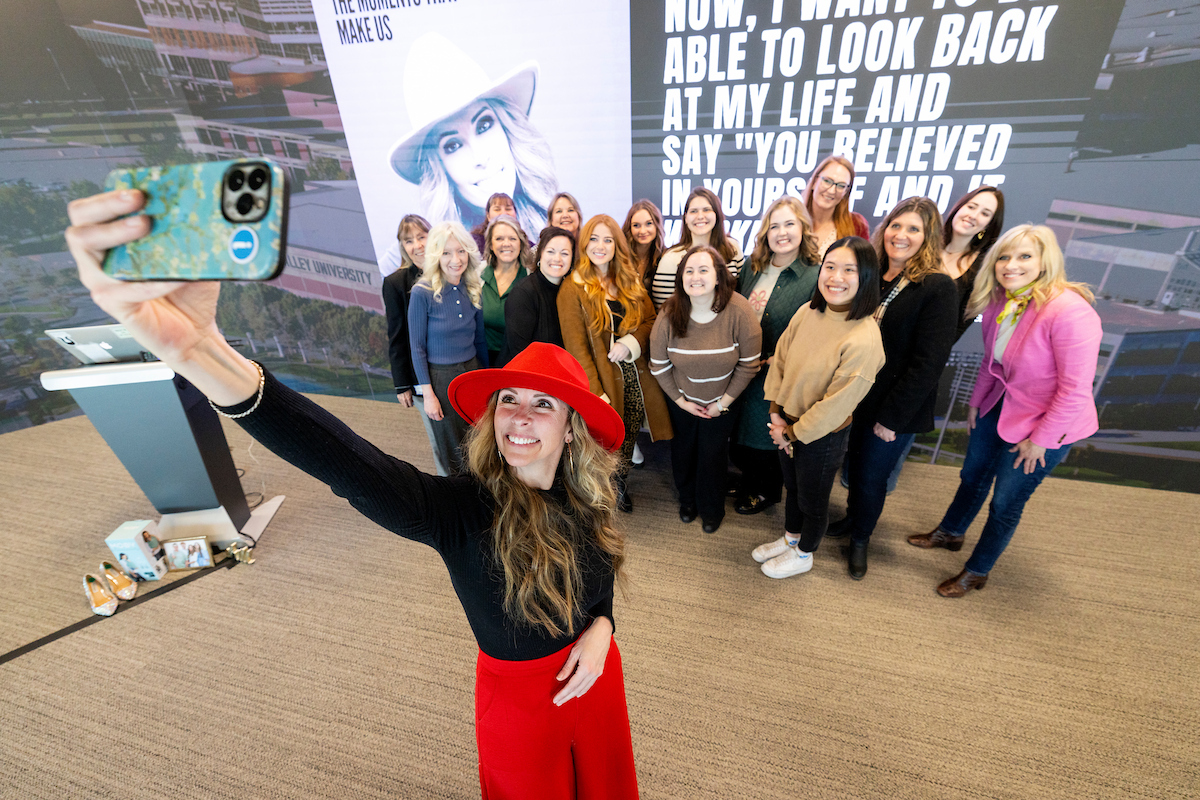 Carrie Hill takes a group selfie with attendees at the Women in Business Impact Lab event, all smiling in front of an inspirational quote backdrop.