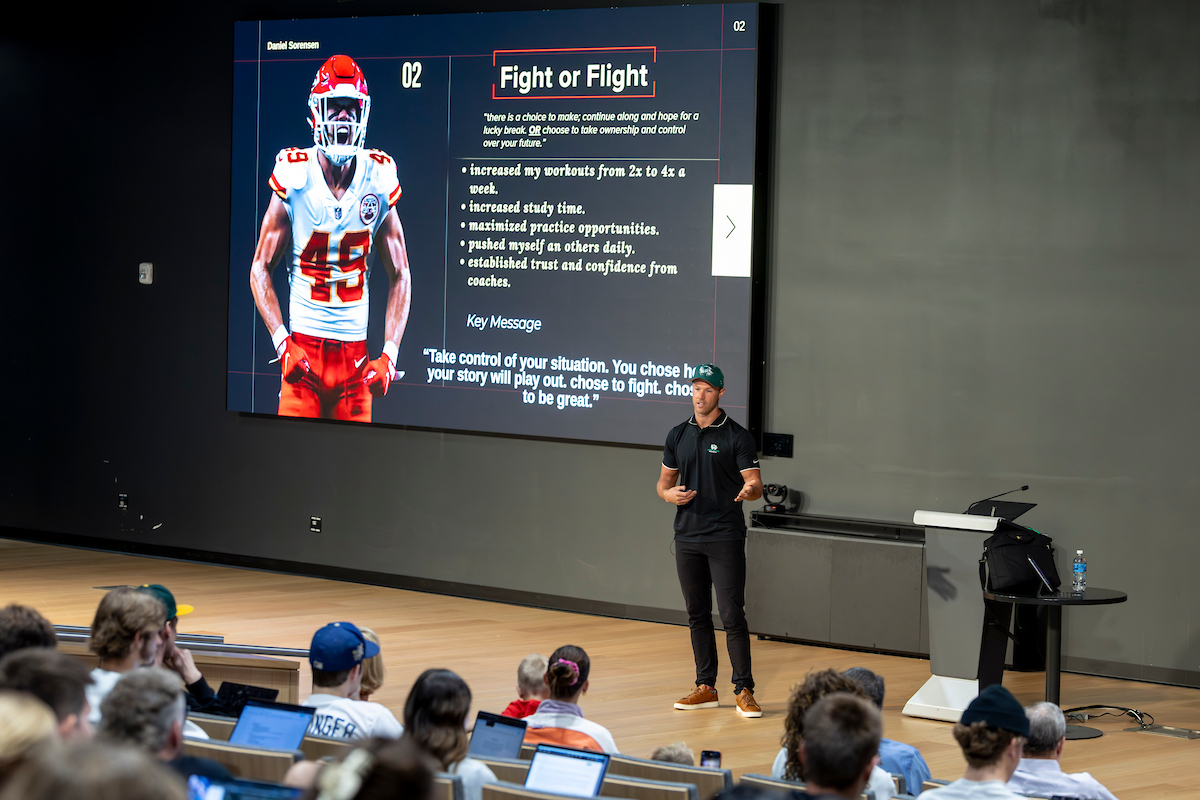 Wide view of Daniel Sorensen addressing UVU students, with a presentation slide titled “Fight or Flight” showing him in his Kansas City Chiefs uniform.