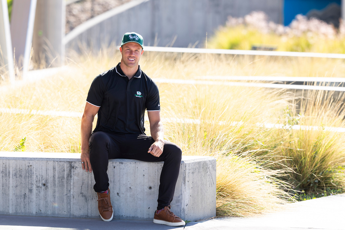 Daniel Sorensen sits outdoors on a concrete bench at UVU, wearing a black polo and green Wolverines hat, smiling in the sunlight.