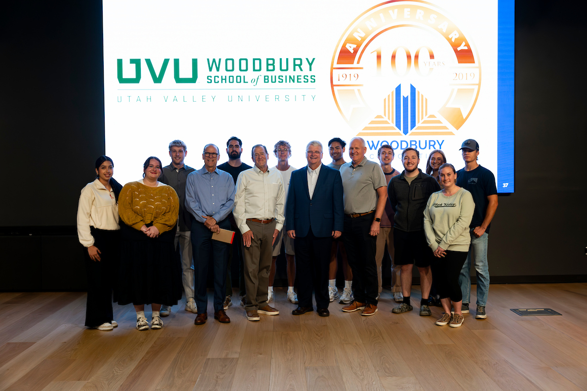 Group photo of Guy Woodbury with UVU students and faculty in front of the Woodbury School of Business and centennial logos following the lecture.