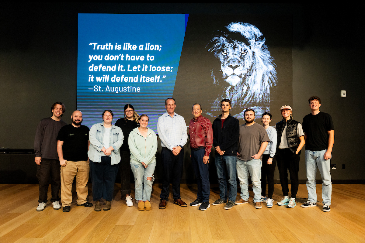 Adam Gale standing with a group of attendees on a stage for a photo. Behind them, a large screen displays a quote: “Truth is like a lion; you don’t have to defend it. Let it loose."