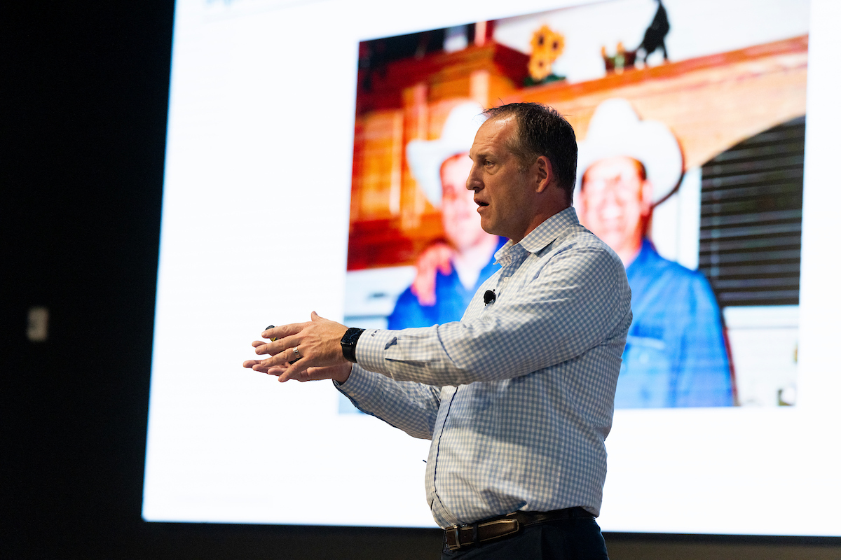 Adam Gale speaks into a microphone while standing in front of a large presentation screen displaying an image of two people wearing cowboy hats.