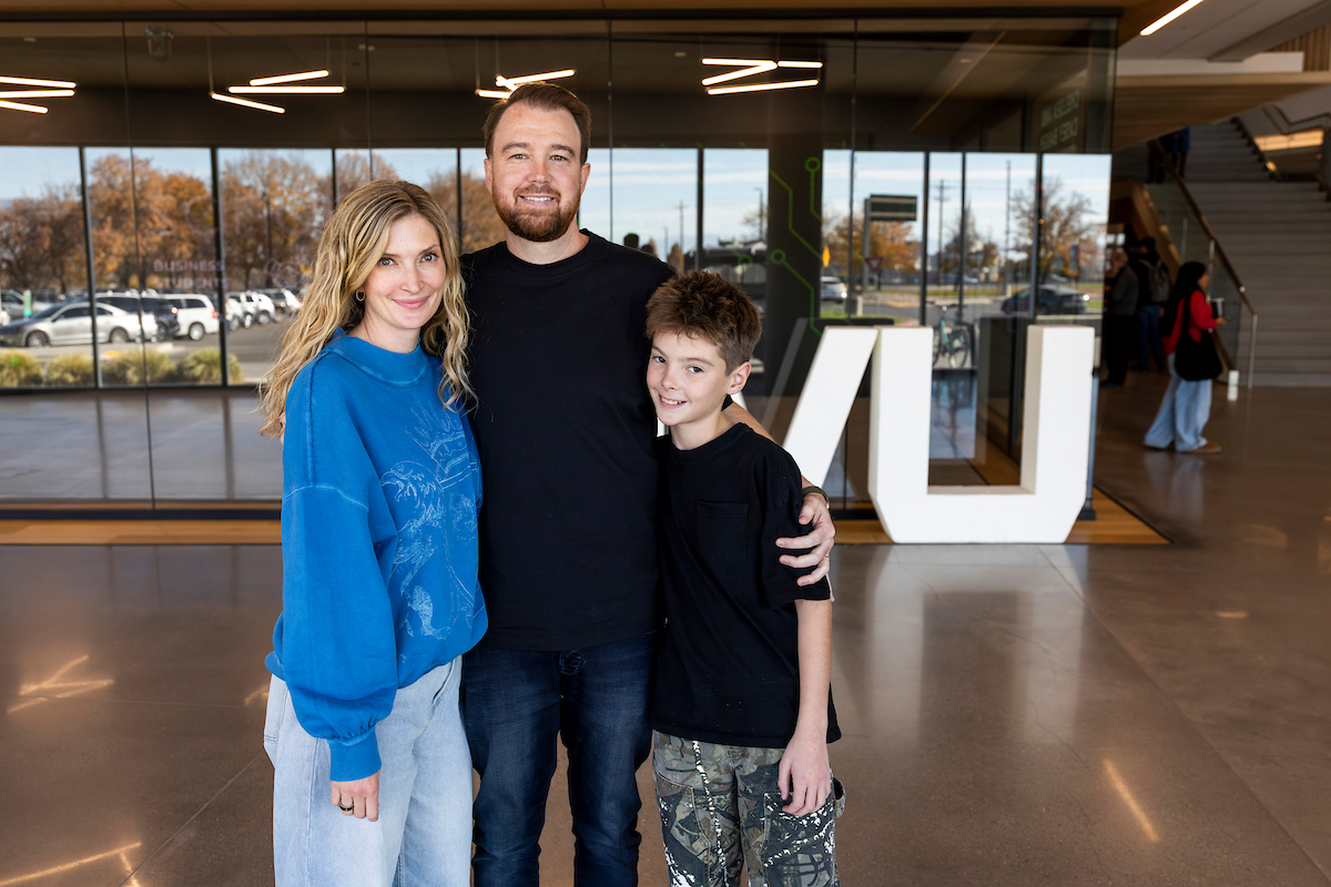 Isaac Westwood stands smiling in a modern lobby area with his family, posing for a photo. Large glass windows and a white UVU sign are visible in the background.