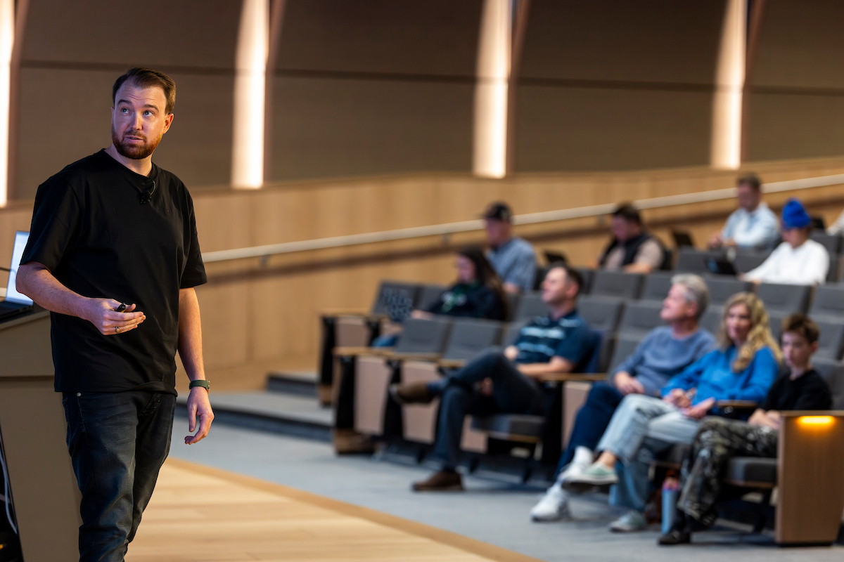 Isaac Westwood stands on stage, holding a presentation remote. Several audience members are seated in gray chairs in the background.