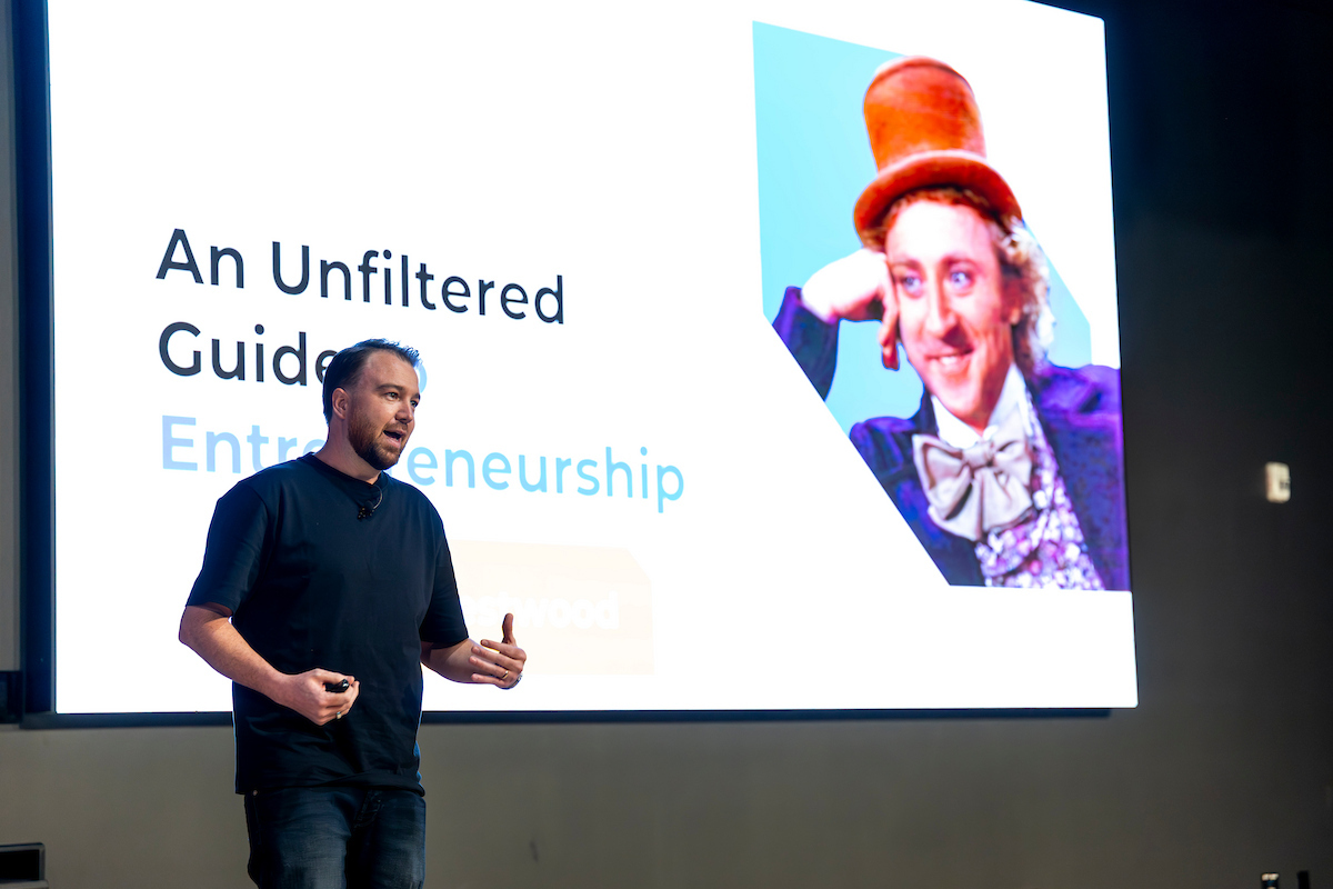 Isaac Westwood presents on stage in front of a large screen displaying the text “An Unfiltered Guide to Entrepreneurship” alongside an image of actor Gene Wilder in Willy Wonka and the Chocolate Factory (1971).
