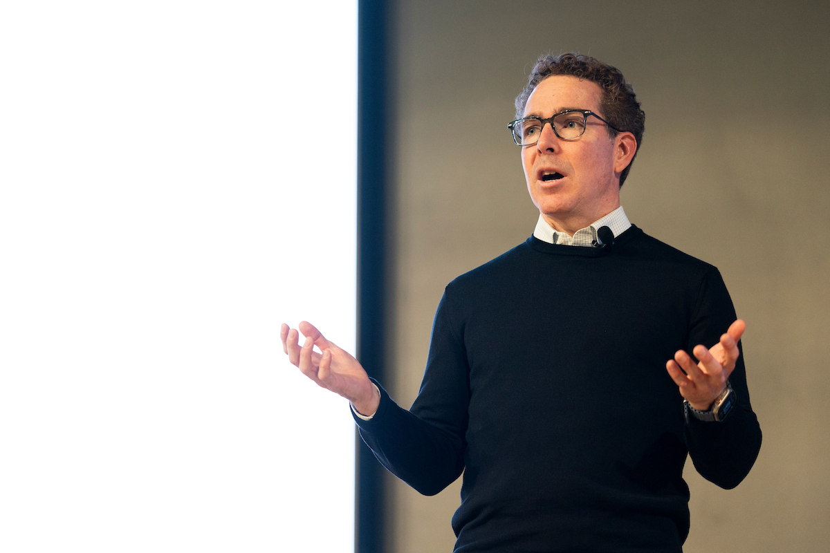 Speaker Mr. Jean Drouin speaks to the audience while standing in front of a projector screen on stage.