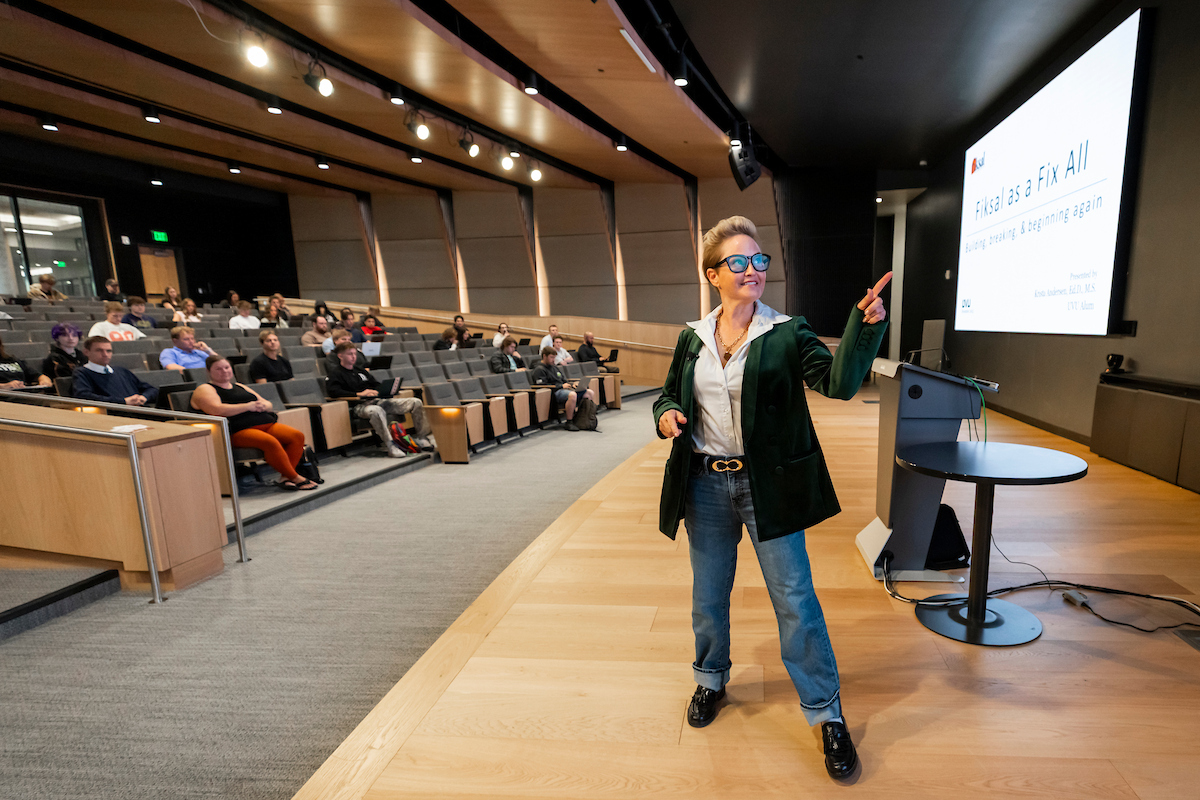 Dr. Krista Anderson stands at the front of a lecture hall, gesturing toward a large presentation screen. The screen displays the title “Fiksal as a Fix All” along with additional text. Several attendees are seated in tiered rows behind the speaker. 