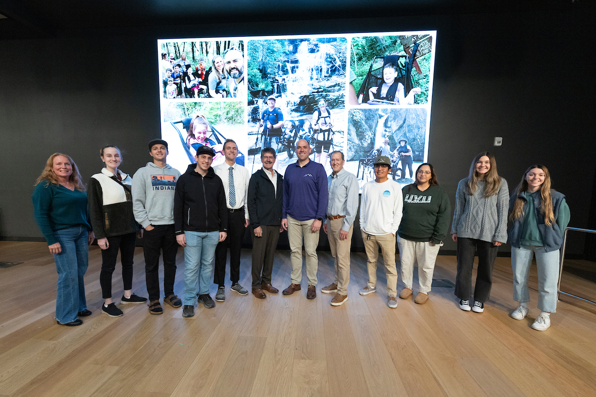  A group photo on stage featuring speaker, LJ Wilde and attendees. Behind them, a large screen displays a collage of outdoor adventure images including waterfalls and hiking scenes. 