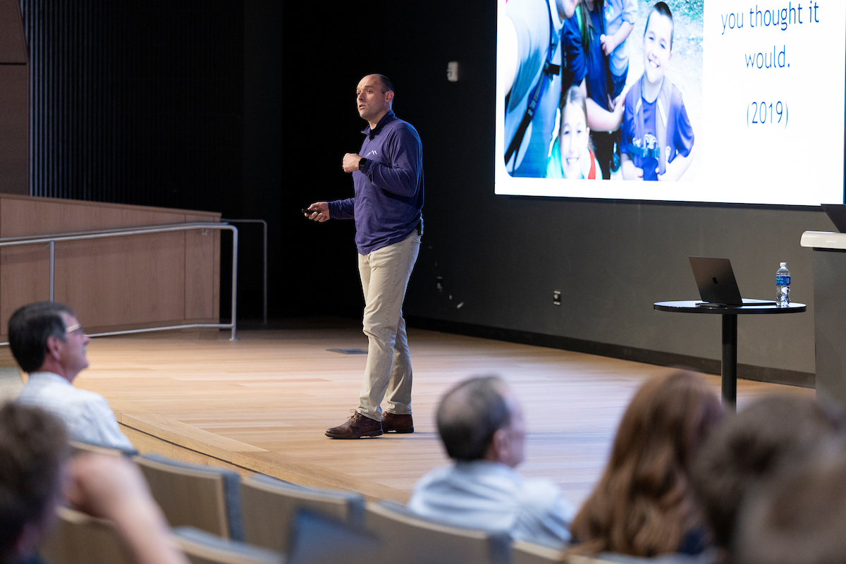 A wide view of the lecture hall showing speaker, LJ Wilde presenting near a large screen with family photos and text. Audience members are seated in tiered rows.