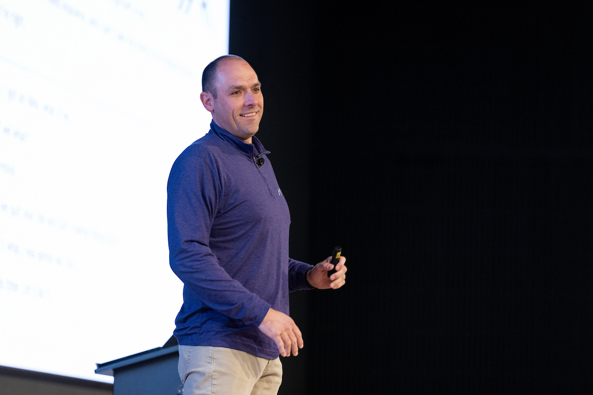 LJ Wilde speaks to an audience on stage in front of a large presentation screen. Wilde is holding a presentation remote and standing near a podium at UVU’s Halladay Lecture Series.
